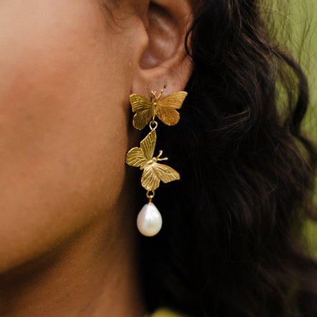 A close-up image of a person's ear wearing a gold-plated earring featuring two butterflies and a pearl.