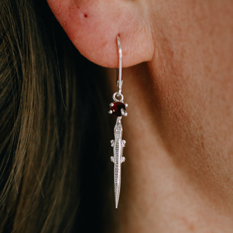 A close-up image of a person's ear wearing a silver hoop earring with an alligator design and a red garnet embellishment.