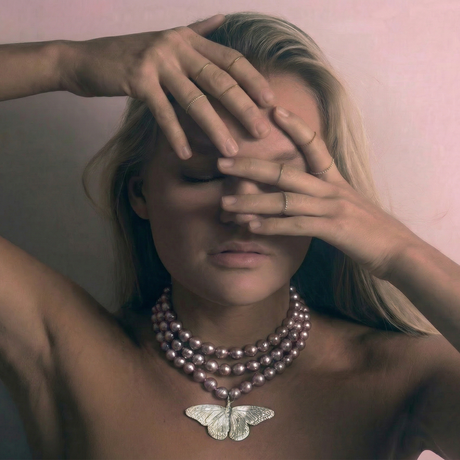 Woman with hands covering face, wearing a butterfly necklace against a pink background